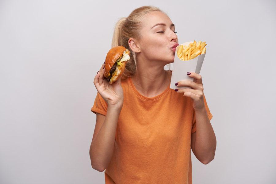 pleased good looking young lovely blonde female casual clothes standing white background with fast food raised hands keeping eyes closed while foretasting yummy meal e1690197499430